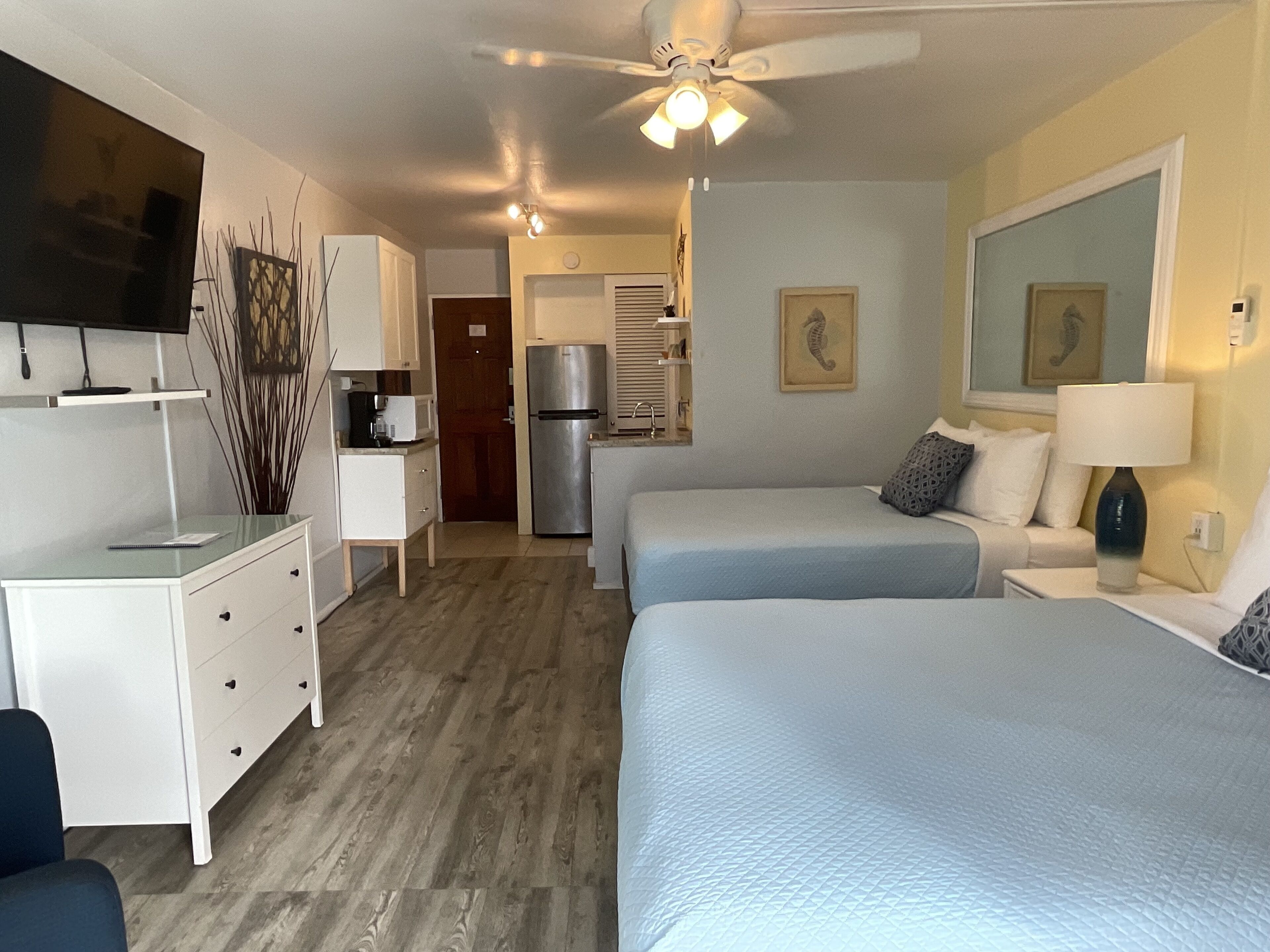Habitación de hotel con dos camas, escritorio, televisor, ventilador de techo y kitchenette.