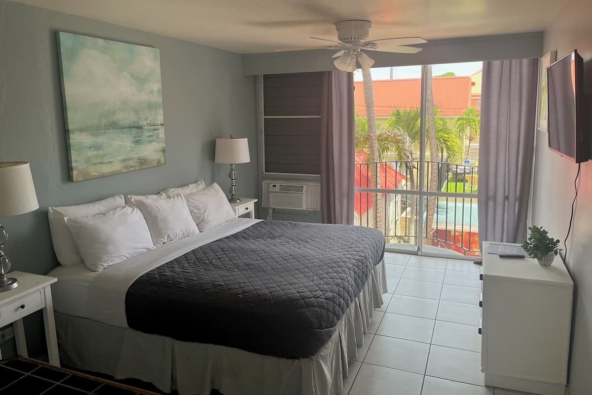 Habitación de hotel con cama, ventilador de techo, un cuadro, ventana con cortinas y vista a una piscina.