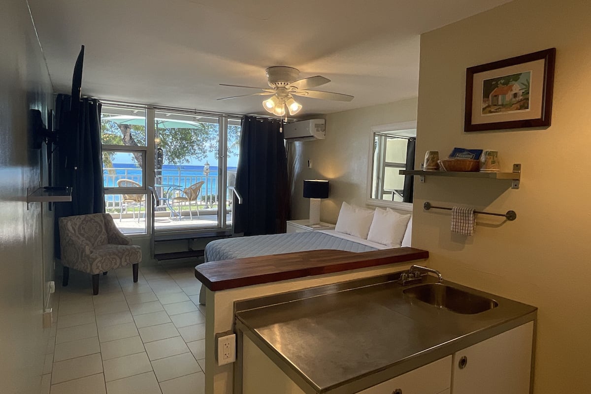 Habitación de hotel con cama, un escritorio pequeño con lavamanos, una silla, ventilador de techo y vista al mar por la ventana.