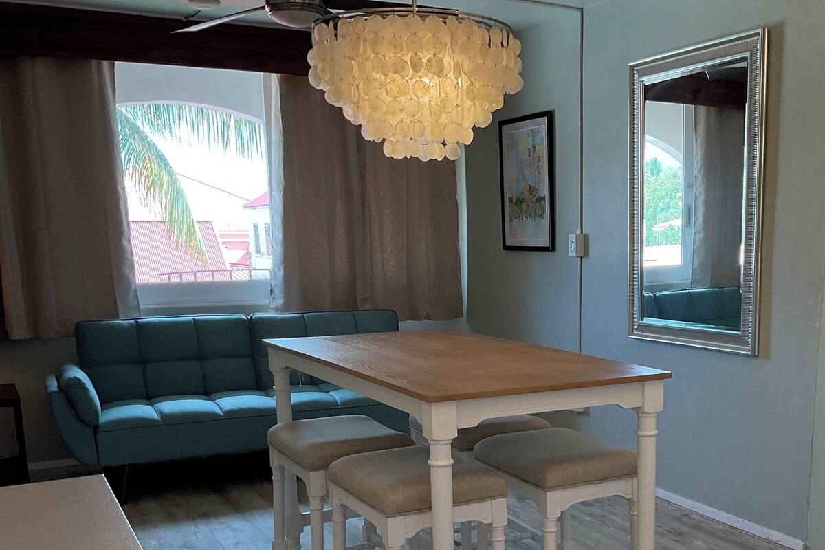 Una habitación con una mesa de comedor de madera, dos sillas blancas, un sofá azul, una araña y un espejo grande.