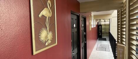 A hallway with a red wall, a framed artwork of a bird and plant, and a fire exit sign.