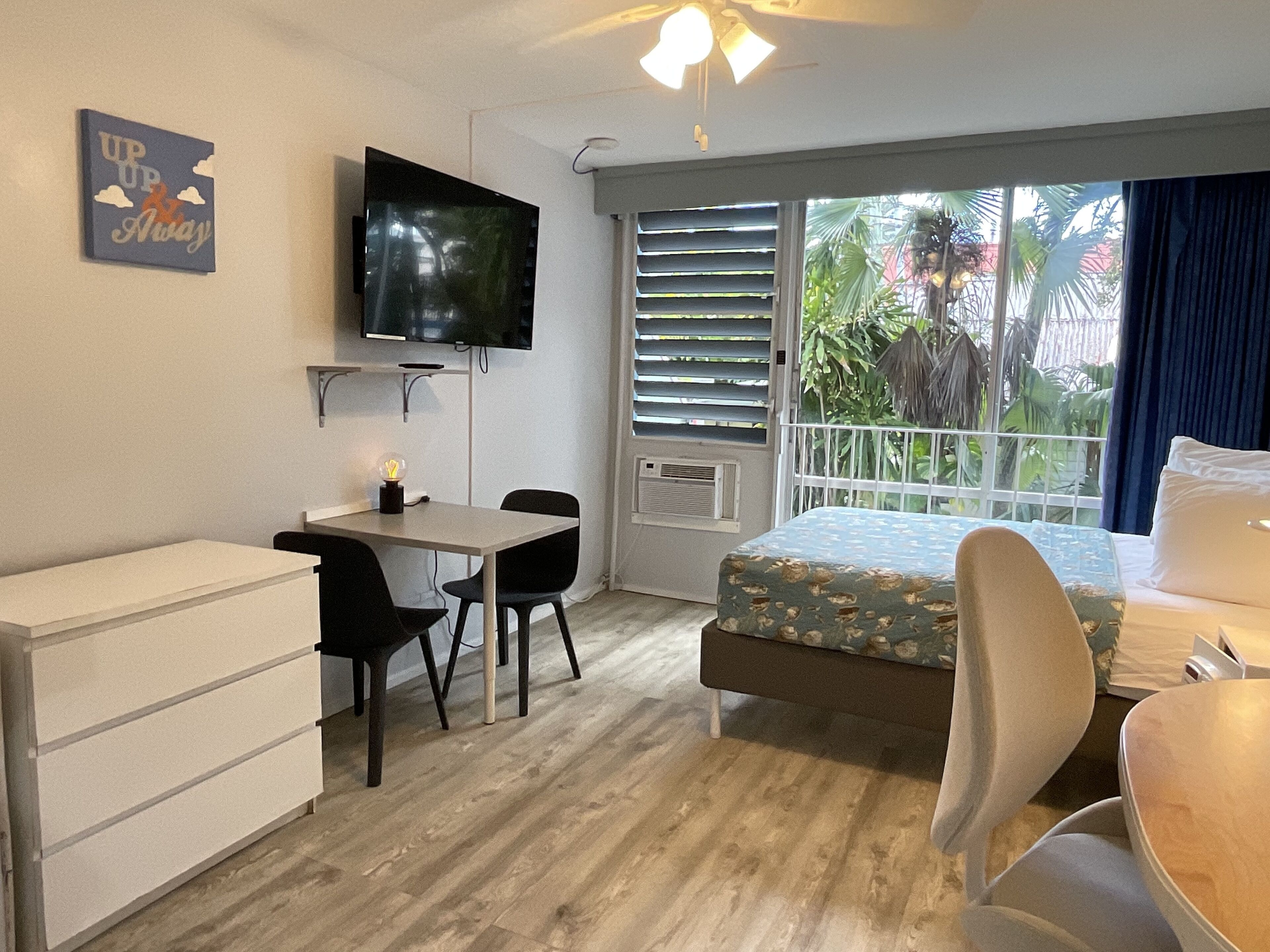 A hotel room with a bed, a desk with a chair, a television, a window with blinds, and a balcony with plants.