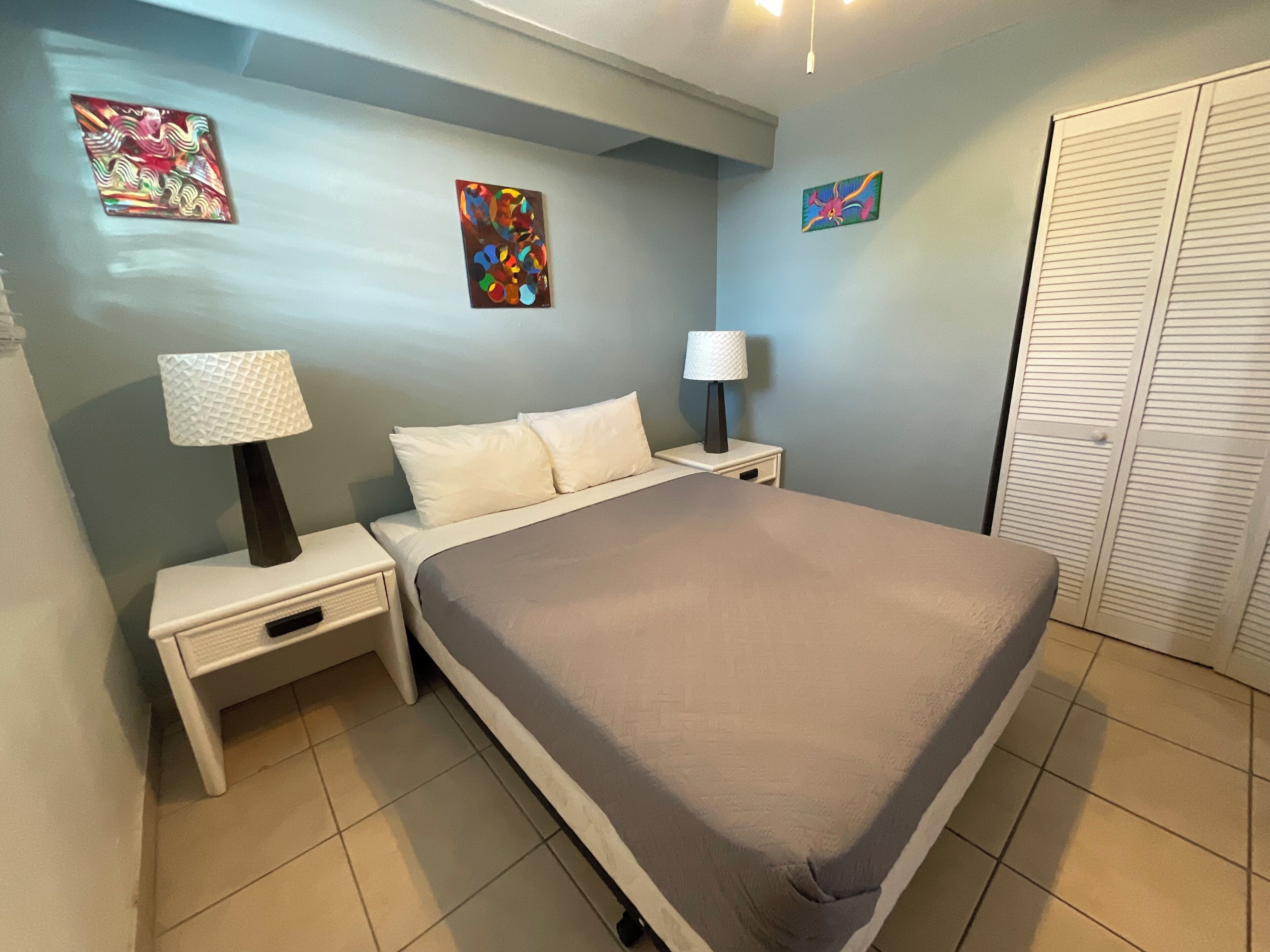 A bedroom with a bed, two bedside tables with lamps, a wardrobe, and two wall art pieces.