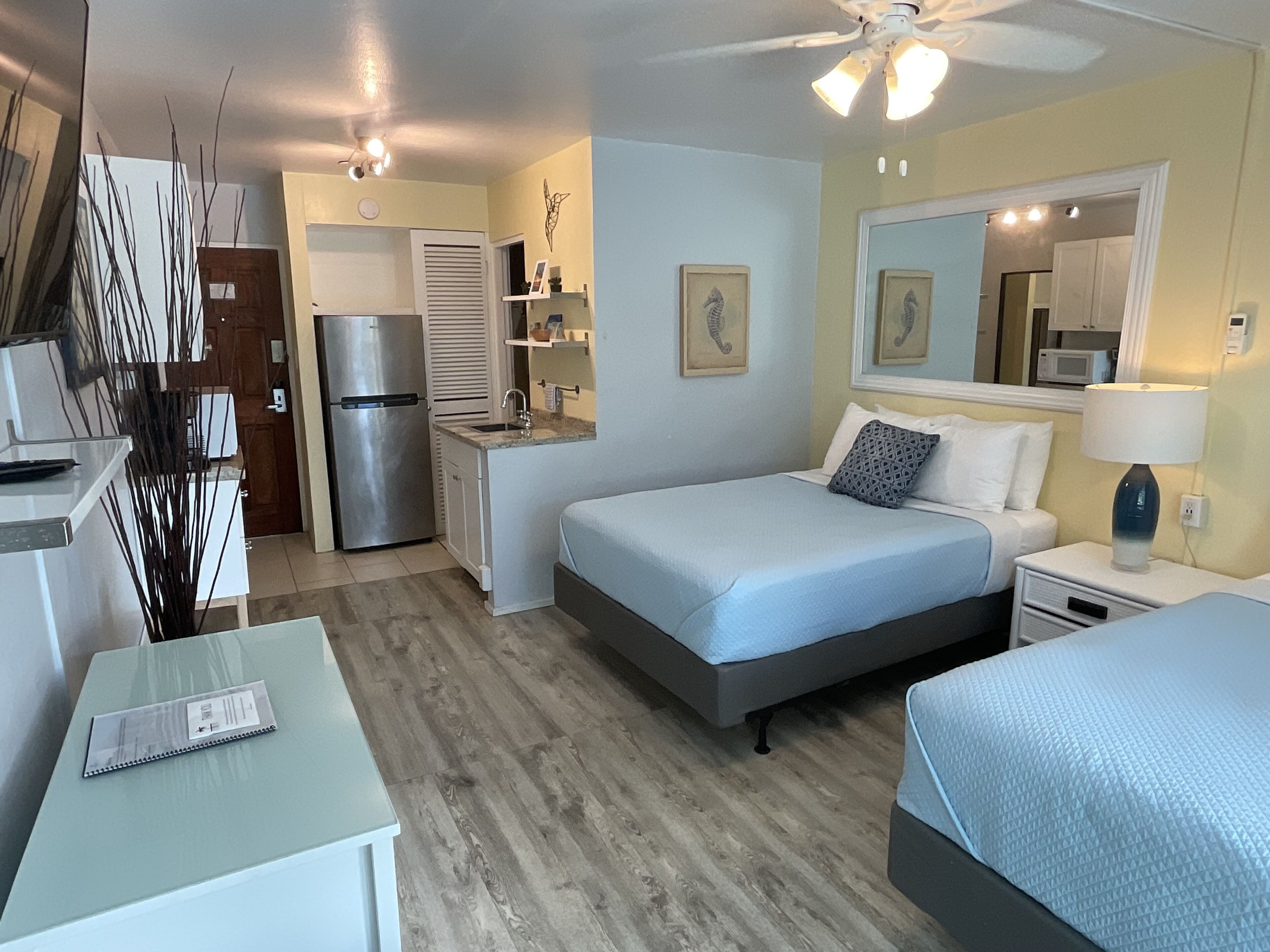 Une chambre d’hôtel compacte comprenant un lit, une table de chevet et un petit bureau avec une chaise. Elle dispose également d’une kitchenette équipée d’un réfrigérateur et d’un four à micro-ondes.