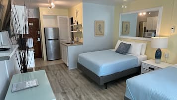 Une chambre d’hôtel compacte comprenant un lit, une table de chevet et un petit bureau avec une chaise. Elle dispose également d’une kitchenette équipée d’un réfrigérateur et d’un four à micro-ondes.