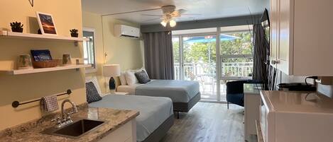 A hotel room with a kitchenette, two beds, a ceiling fan, a large window with a view, and a balcony with chairs.