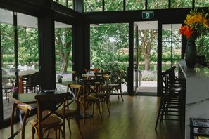 Un comedor interior con mesas y sillas de madera, ventanas amplias que ofrecen vista a un jardín con árboles y flores.