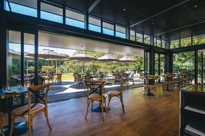 Un comedor interior con sillas y mesas de madera, ventanas amplias y sombrillas exteriores.