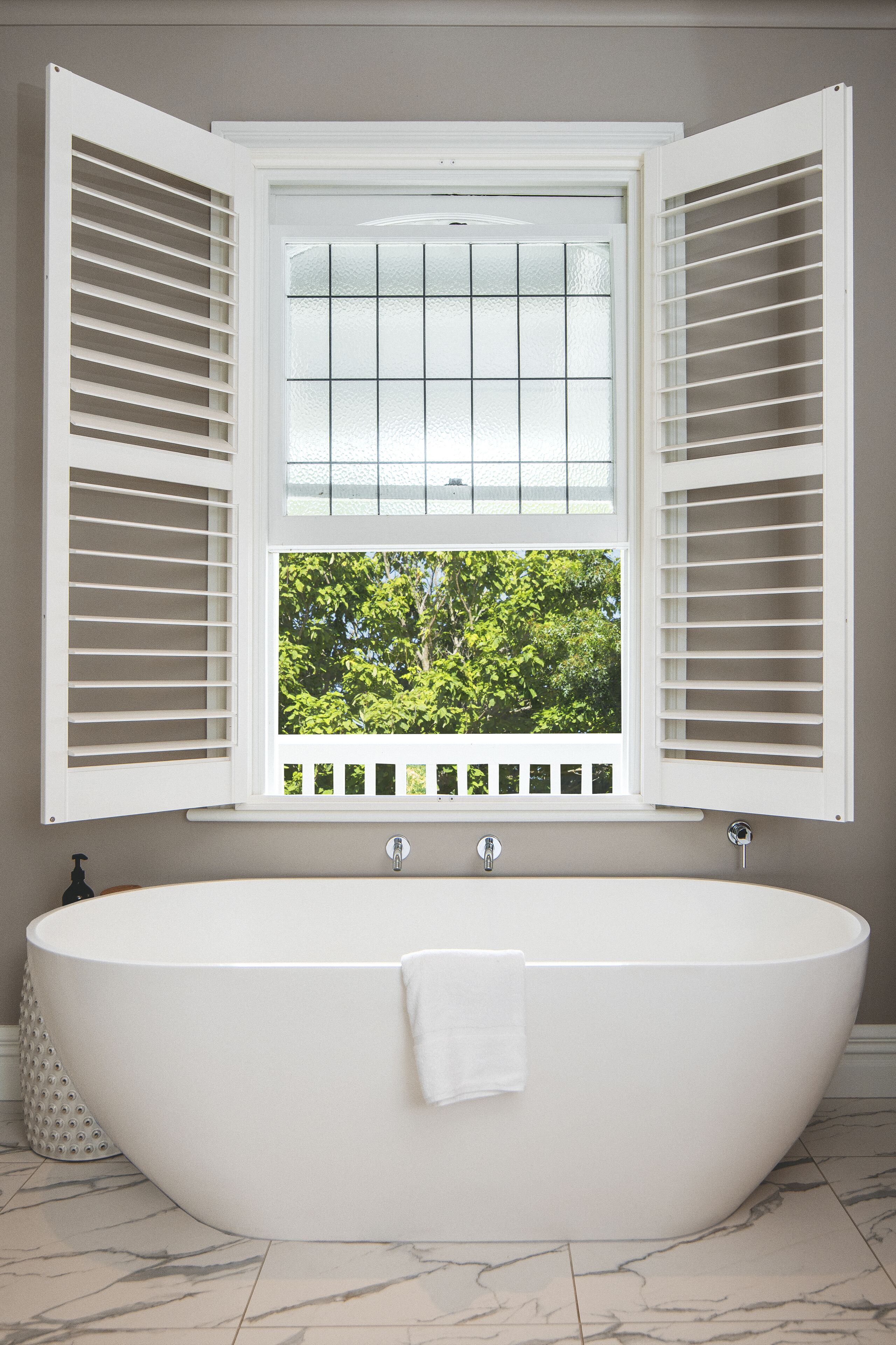A modern bathroom with a freestanding bathtub, a window with shutters, and a view of greenery outside.
