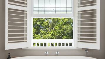 A modern bathroom with a freestanding bathtub, a window with shutters, and a view of greenery outside.