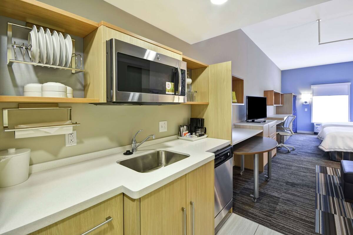 A modern kitchen with a microwave, sink, and dishwasher, a small dining area with a table and chairs, and a bedroom with a bed and window.