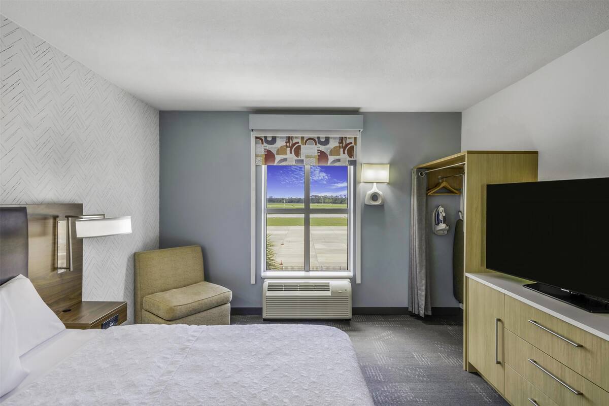 A hotel room with a bed, a TV, a chair, a wardrobe, a window with a view of a runway, and a lamp.