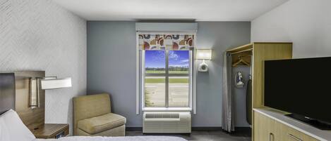 A hotel room with a bed, a TV, a chair, a wardrobe, a window with a view of a runway, and a lamp.