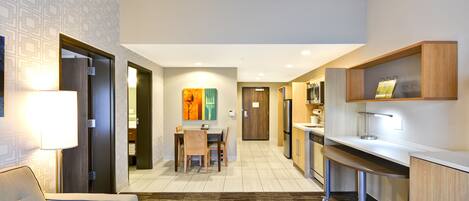 A compact hotel room with a kitchenette, a dining area, and a living space with a sofa and lamp.