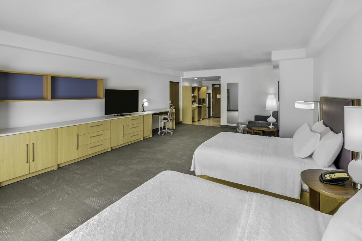 A hotel room with two beds, a desk, a television, and a kitchenette.