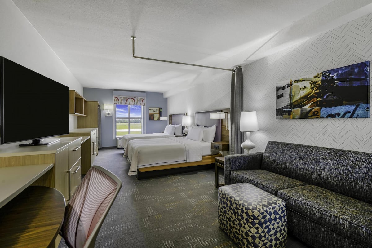 A hotel room with a bed, a desk with a TV, a sofa, a patterned ottoman, and a painting on the wall.