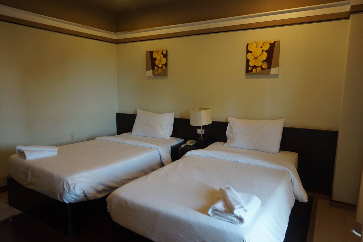 A hotel room with two single beds, each with white linens and pillows, a bedside table with a lamp, and two wall-mounted artworks.