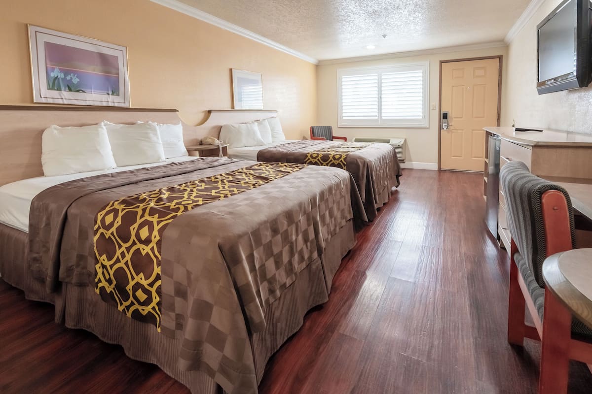 A hotel room with two beds, a TV, a desk, and a window with blinds.