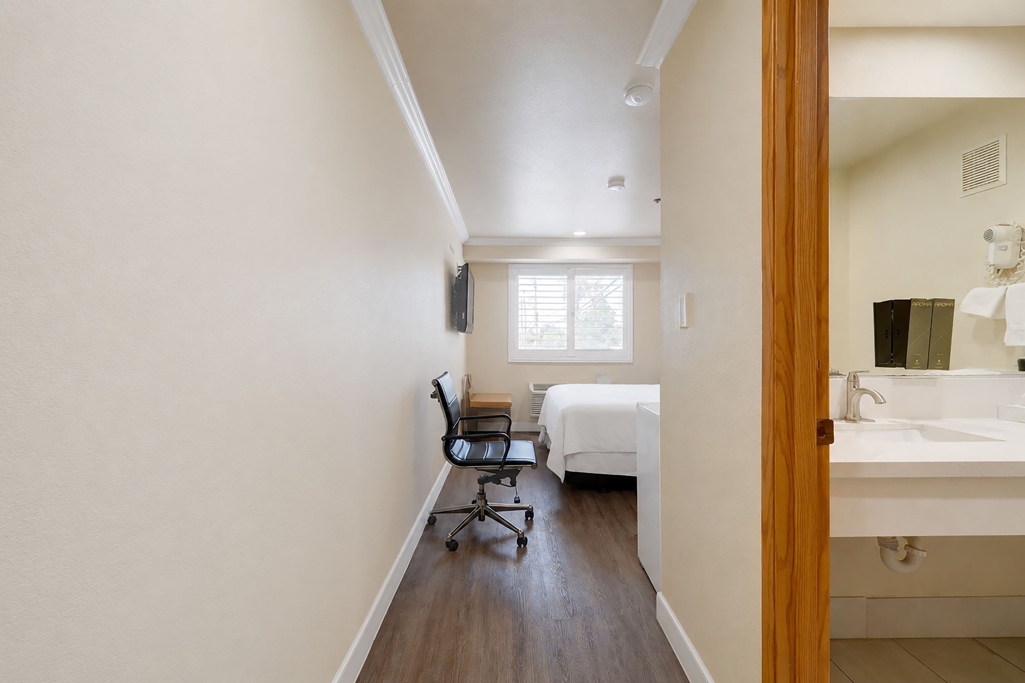 Habitación de hotel con cama, escritorio y silla, ventana con persianas, y baño con lavamanos y espejo.