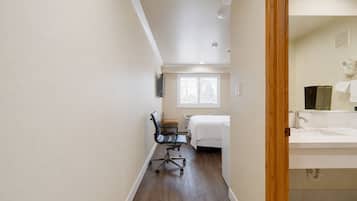 Habitación de hotel con cama, escritorio y silla, ventana con persianas, y baño con lavamanos y espejo.