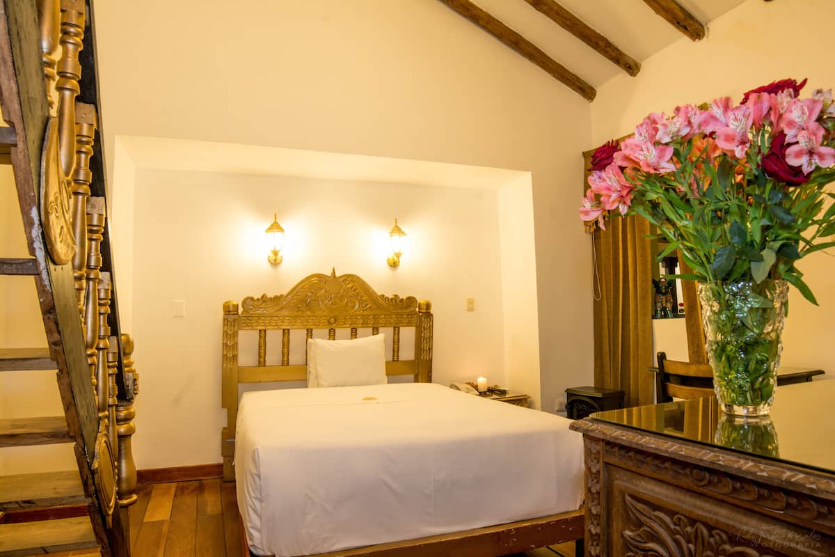 A bedroom with a wooden bed, two wall-mounted lights, a bedside table with a lamp, and a vase with flowers.