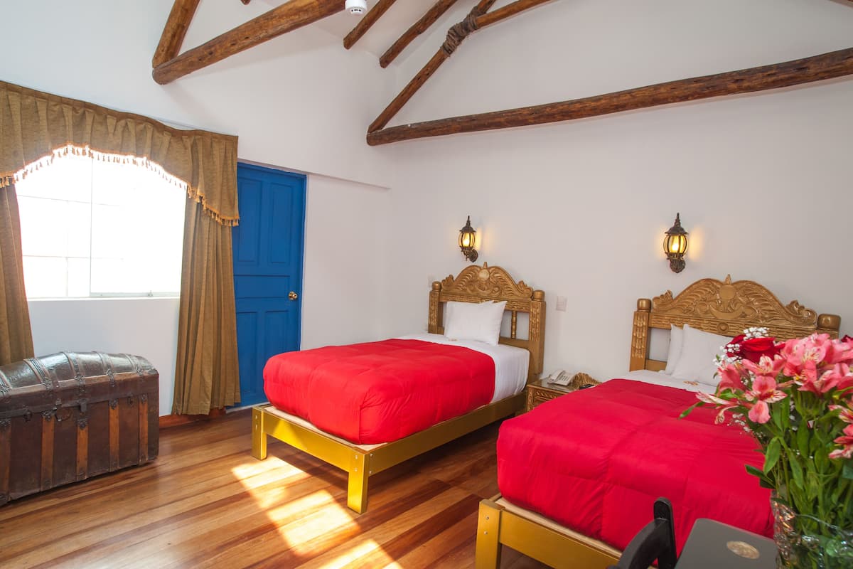 A room with two wooden beds with red bedding, a blue door, a wooden chest, and a vase with flowers.