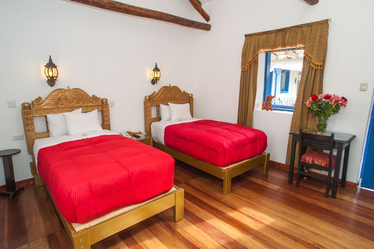 A room with two wooden beds, each with a red bedspread and white pillows. There are two wall-mounted light fixtures, a small round table, and a chair with a red cushion. A vase with flowers is on a small table next to the window.