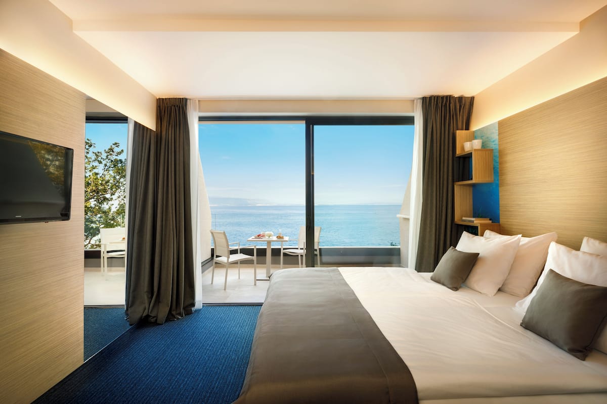 A hotel room with a large bed, a flat-screen TV, a balcony with a table and chairs, and a view of the sea.