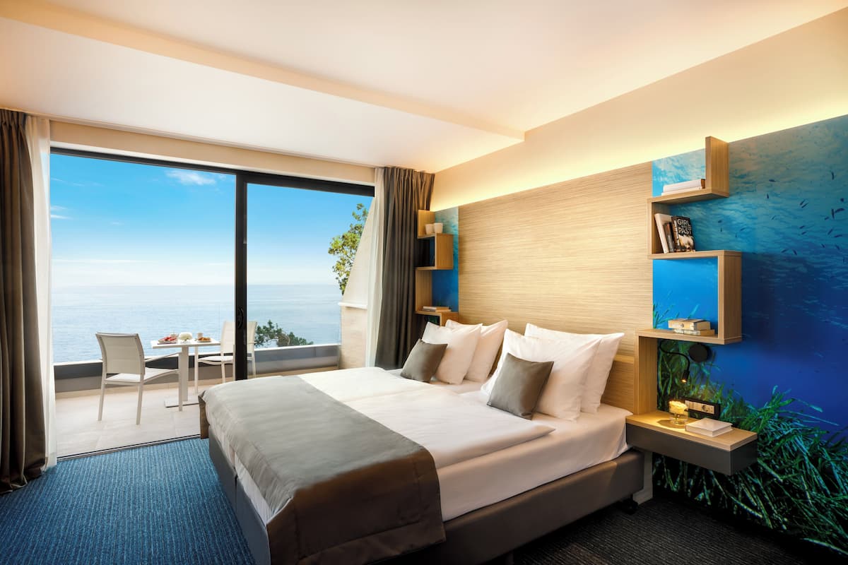 A hotel room with a large bed, a desk with a chair, a view of the sea, and a blue wall with shelves.