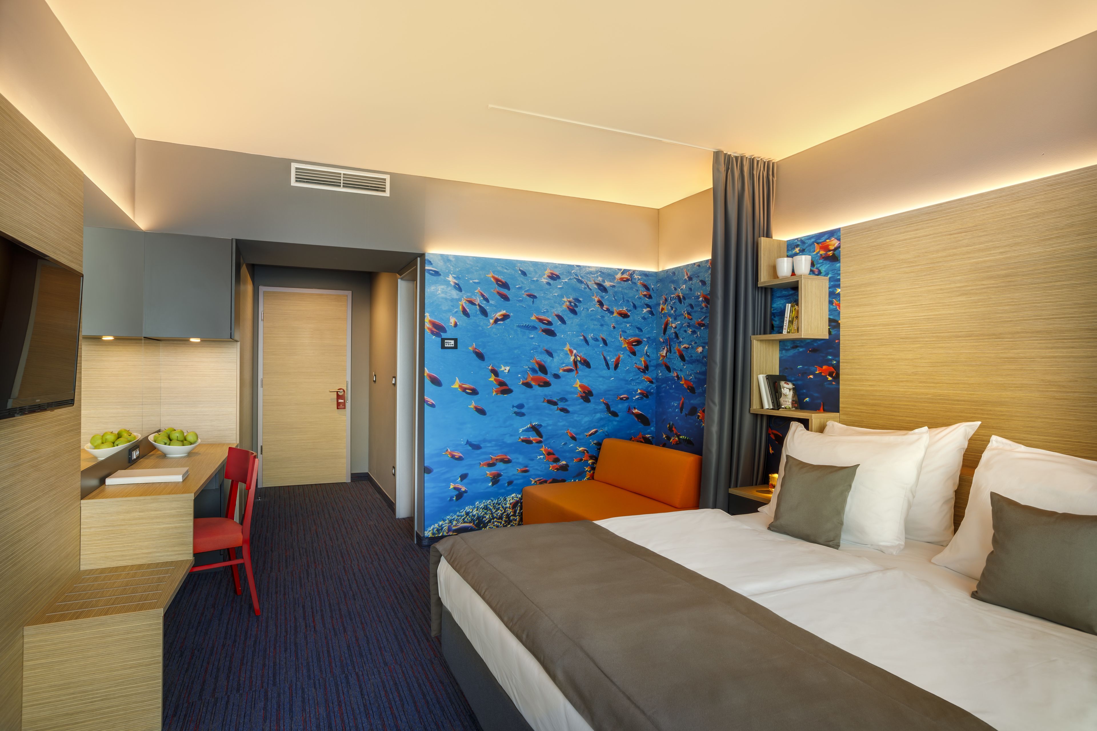 Habitación de hotel con cama, escritorio con silla, estantería y un mural de peces en la pared.