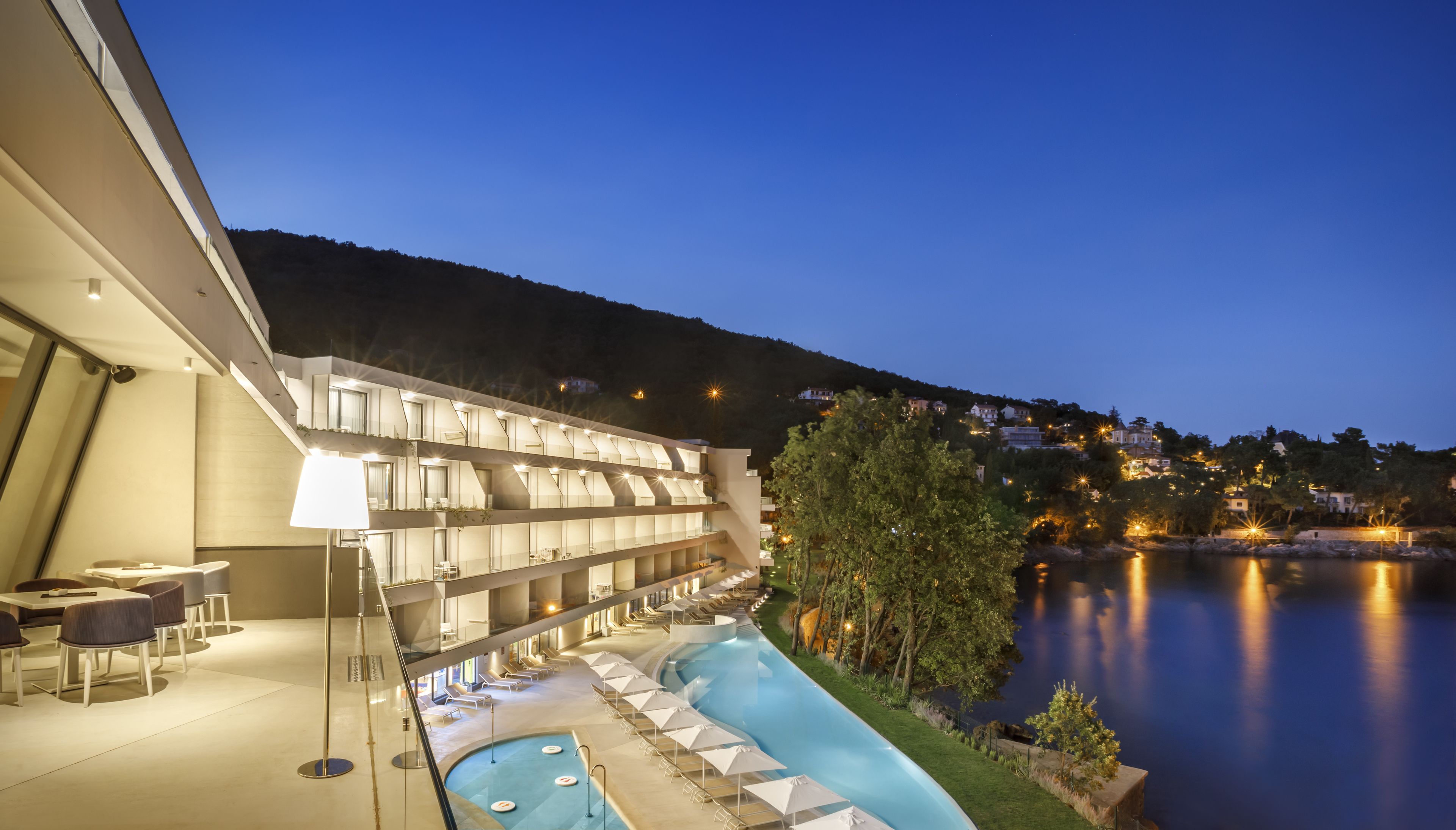 A modern hotel with a pool, outdoor seating, and a view of a lake at dusk.