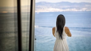 Una mujer con un vestido blanco de pie junto a un ventanal con vista a un cuerpo de agua y montañas a lo lejos.