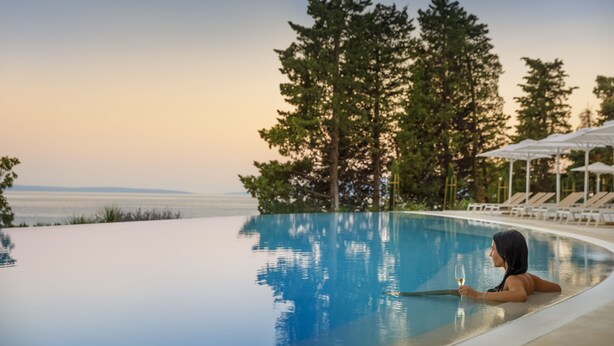 Una persona relajándose en una piscina infinita transparente con vistas al océano y árboles.