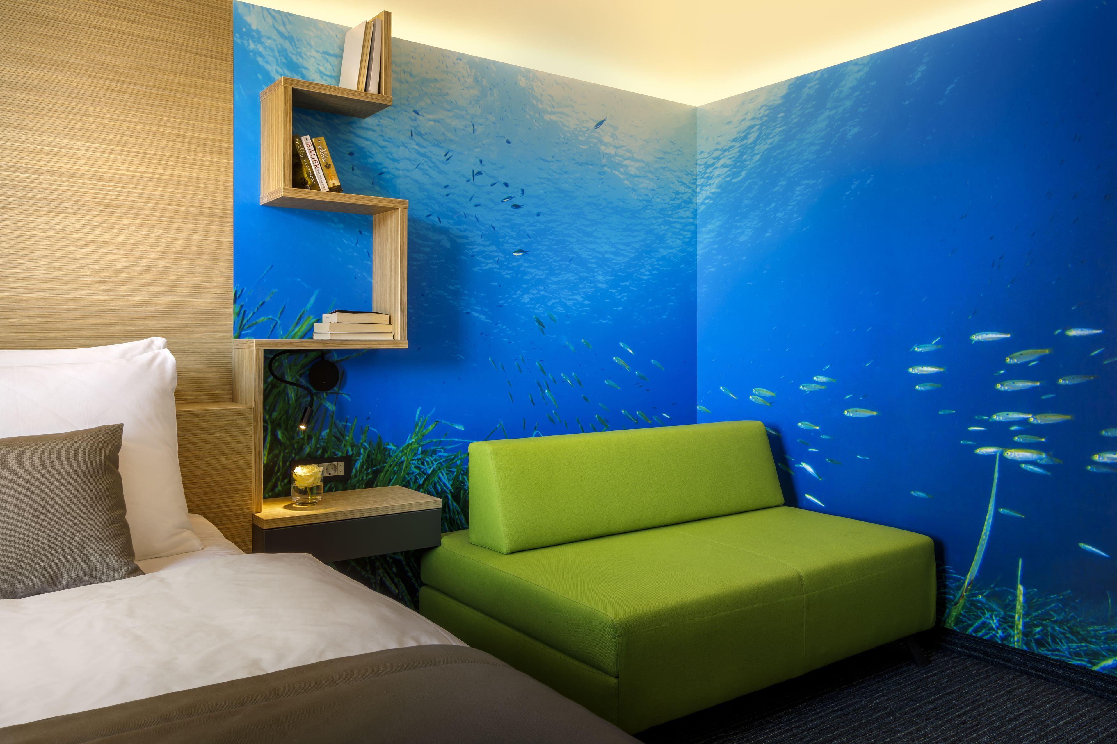 Ein Zimmer mit einem Bett, einem grünen Sofa, einem Nachttisch mit Lampe und einem Wandbild mit Unterwasserlandschaft.