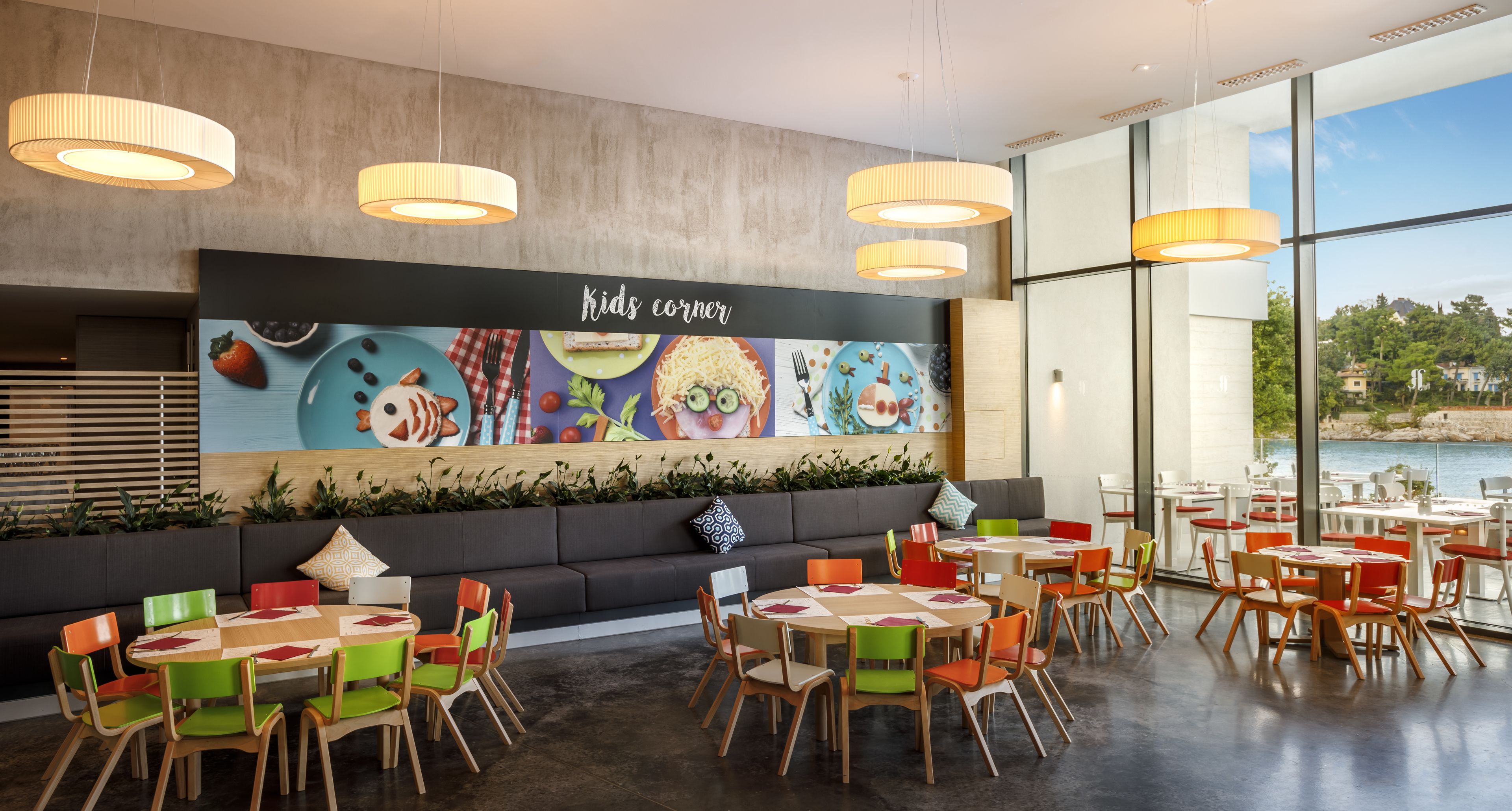 A children's dining area with colorful chairs and tables, a mural with food illustrations, and a view of the sea through large windows.