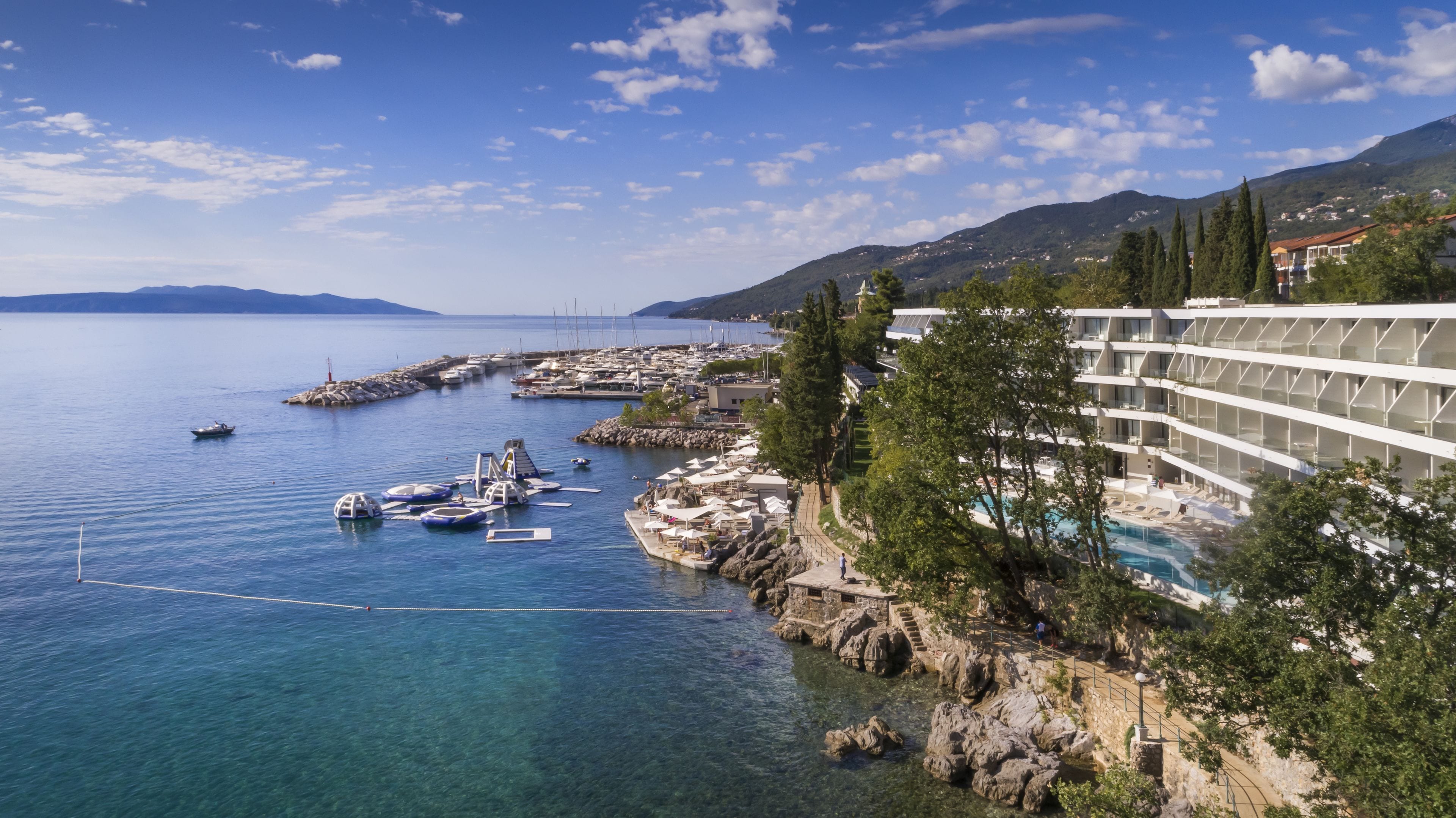 A coastal hotel with a pool, rocky shoreline, and boats in the water.