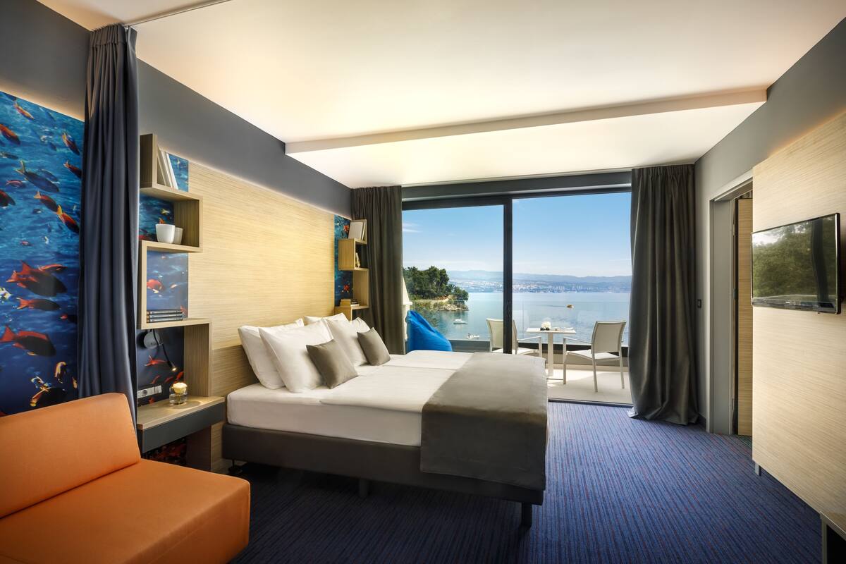 A modern hotel room with a large bed, a desk, and a view of the sea.