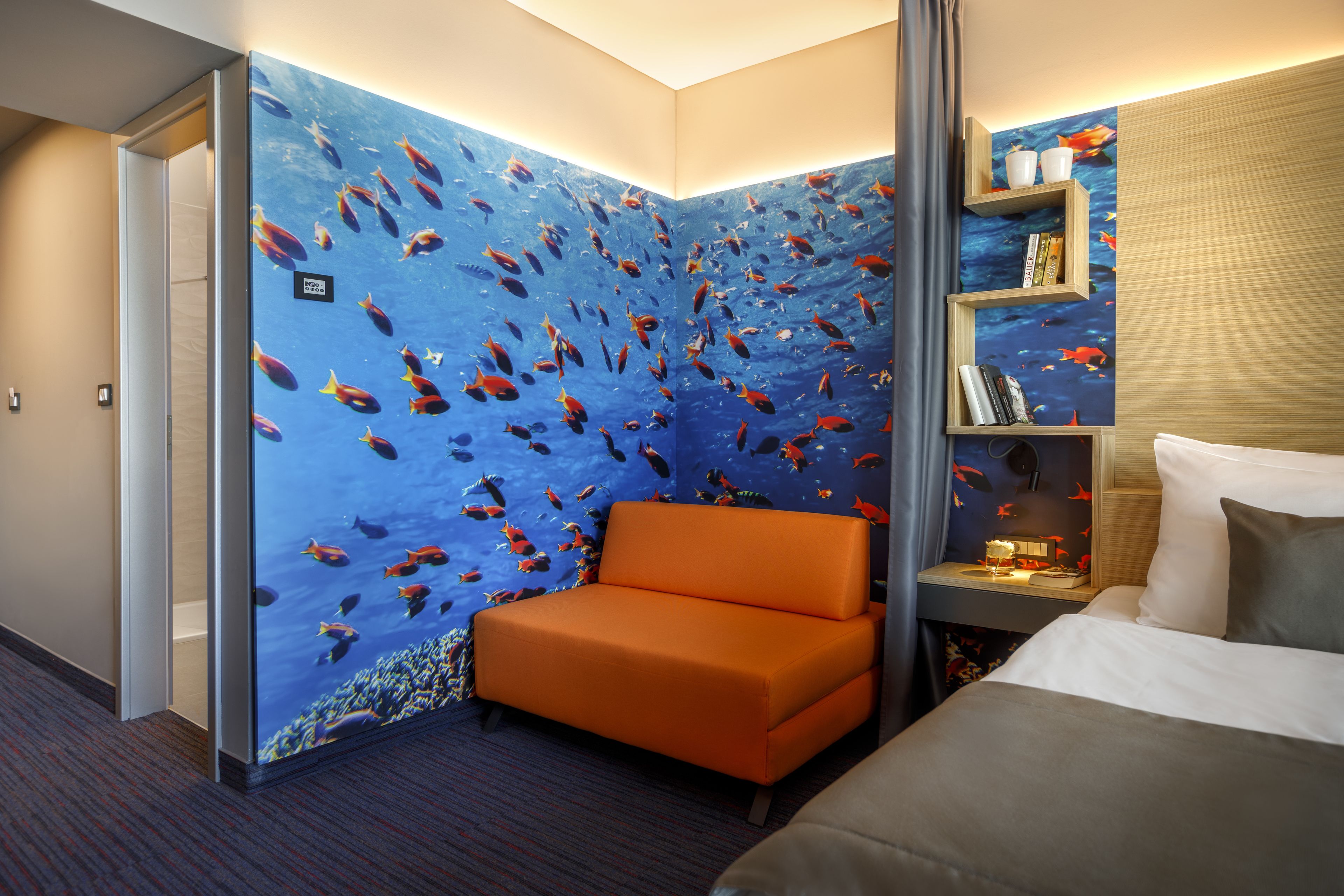Una habitación de hotel con una gran pared tipo acuario, un sofá, una cama y una mesita de noche con libros y una lámpara.