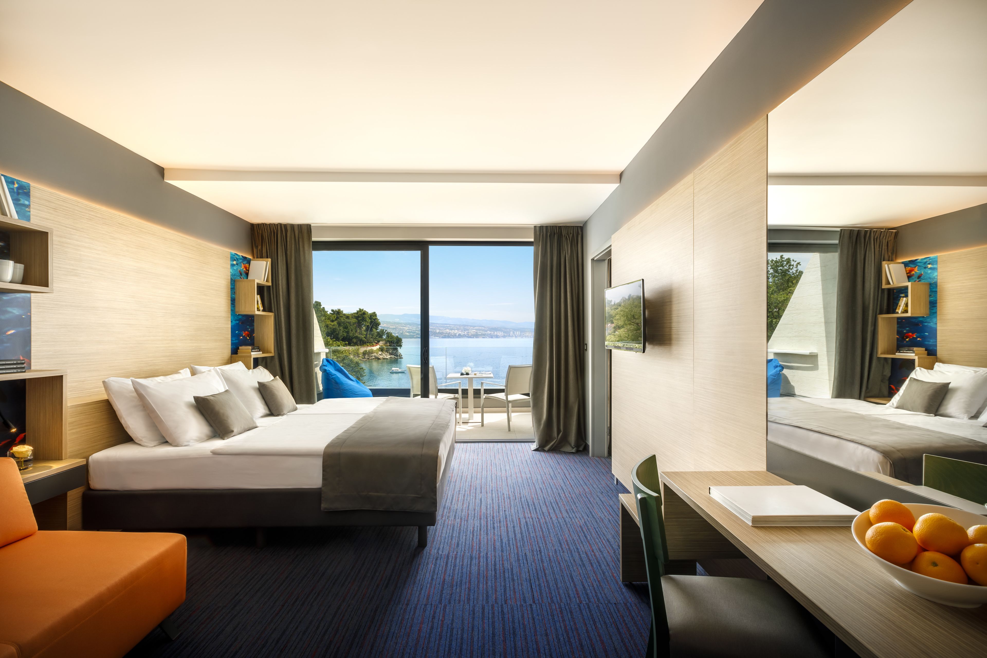 Ett hotellrum med en säng, ett skrivbord, en stol, en soffa, en TV och utsikt över havet.