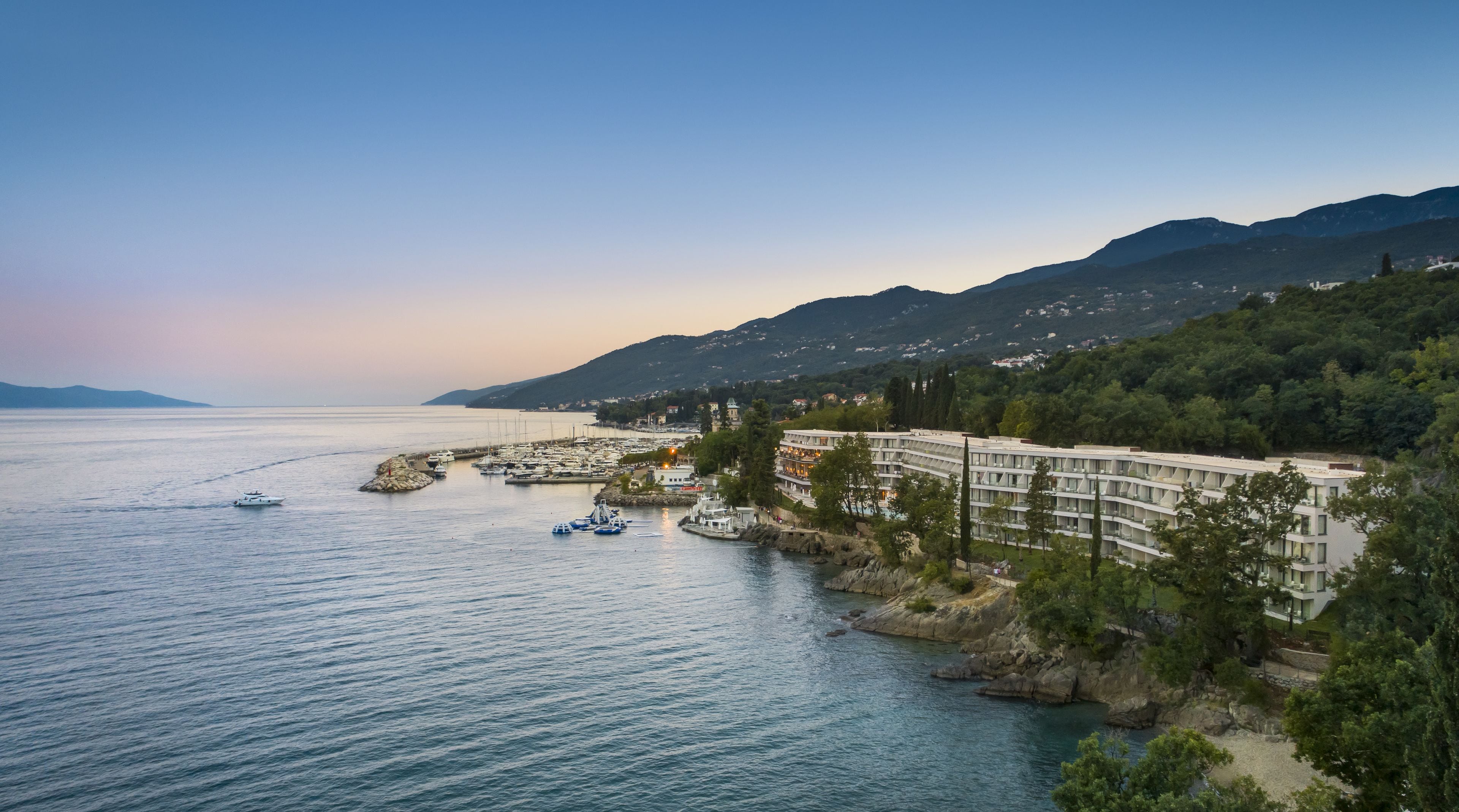 A coastal hotel with multiple balconies overlooking the sea, a marina with boats, and a backdrop of mountains.