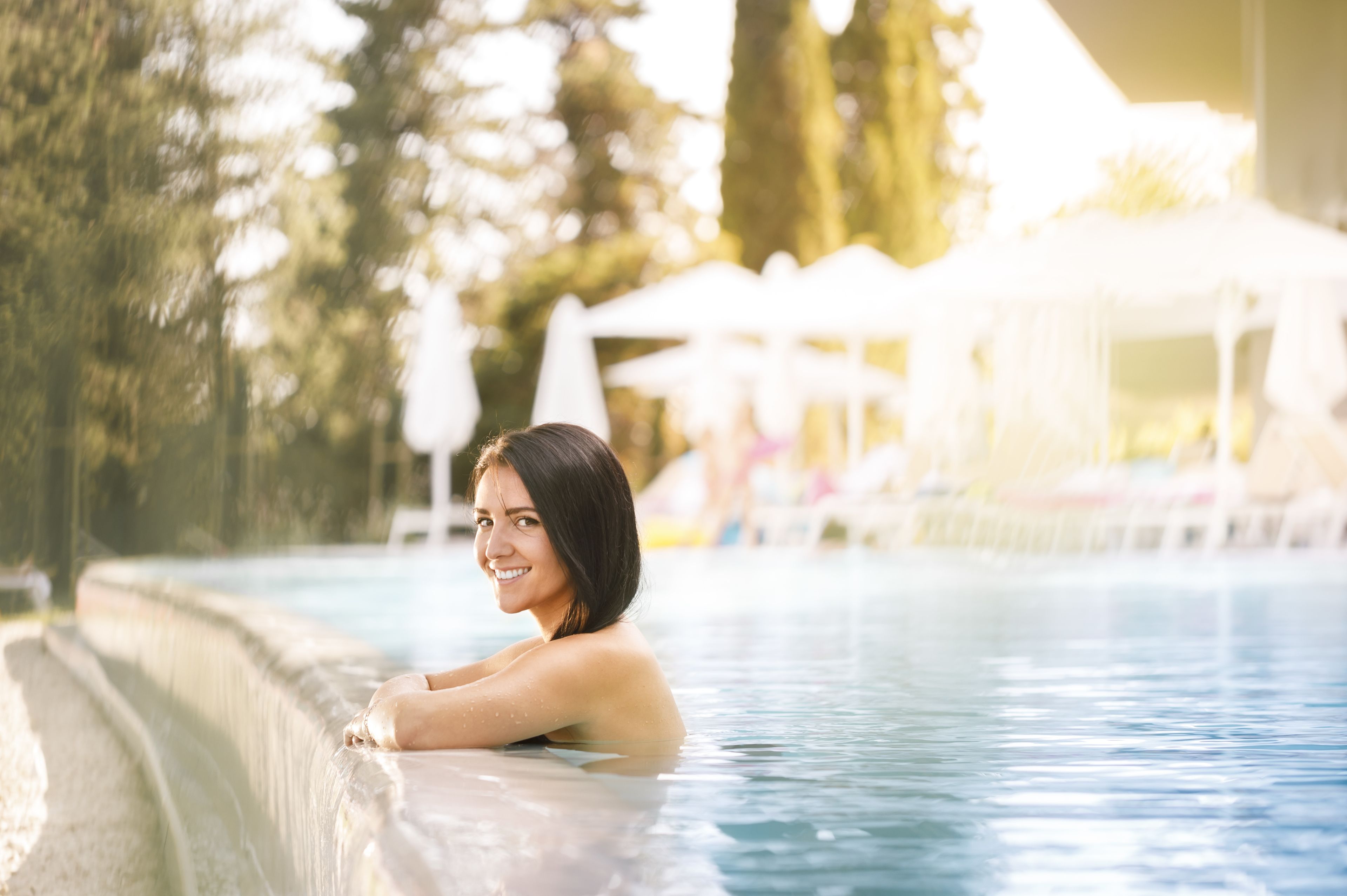 A person relaxing in a swimming pool with a poolside area, umbrellas, and lounge chairs in the background.