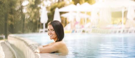 A person relaxing in a swimming pool with a poolside area, umbrellas, and lounge chairs in the background.