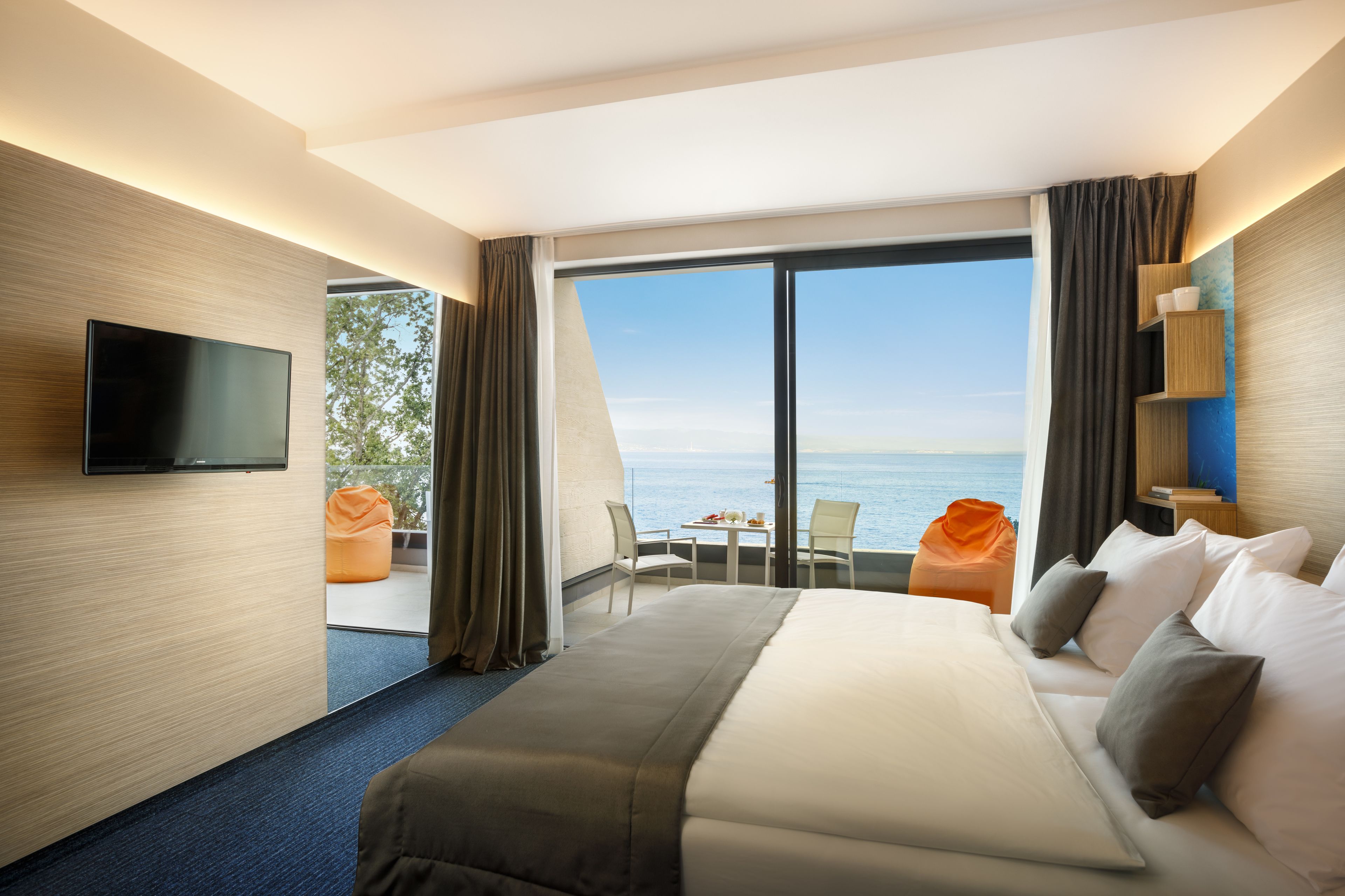 A modern hotel room with a large bed, a flat-screen TV, a dining area with a table and chairs, and a view of the sea.