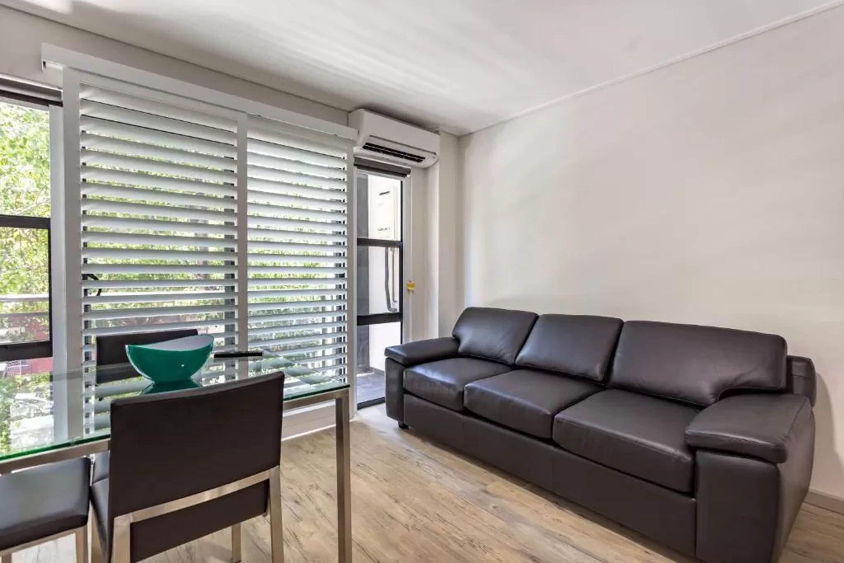 A modern living room with a glass dining table, a brown leather sofa, and a window with blinds.