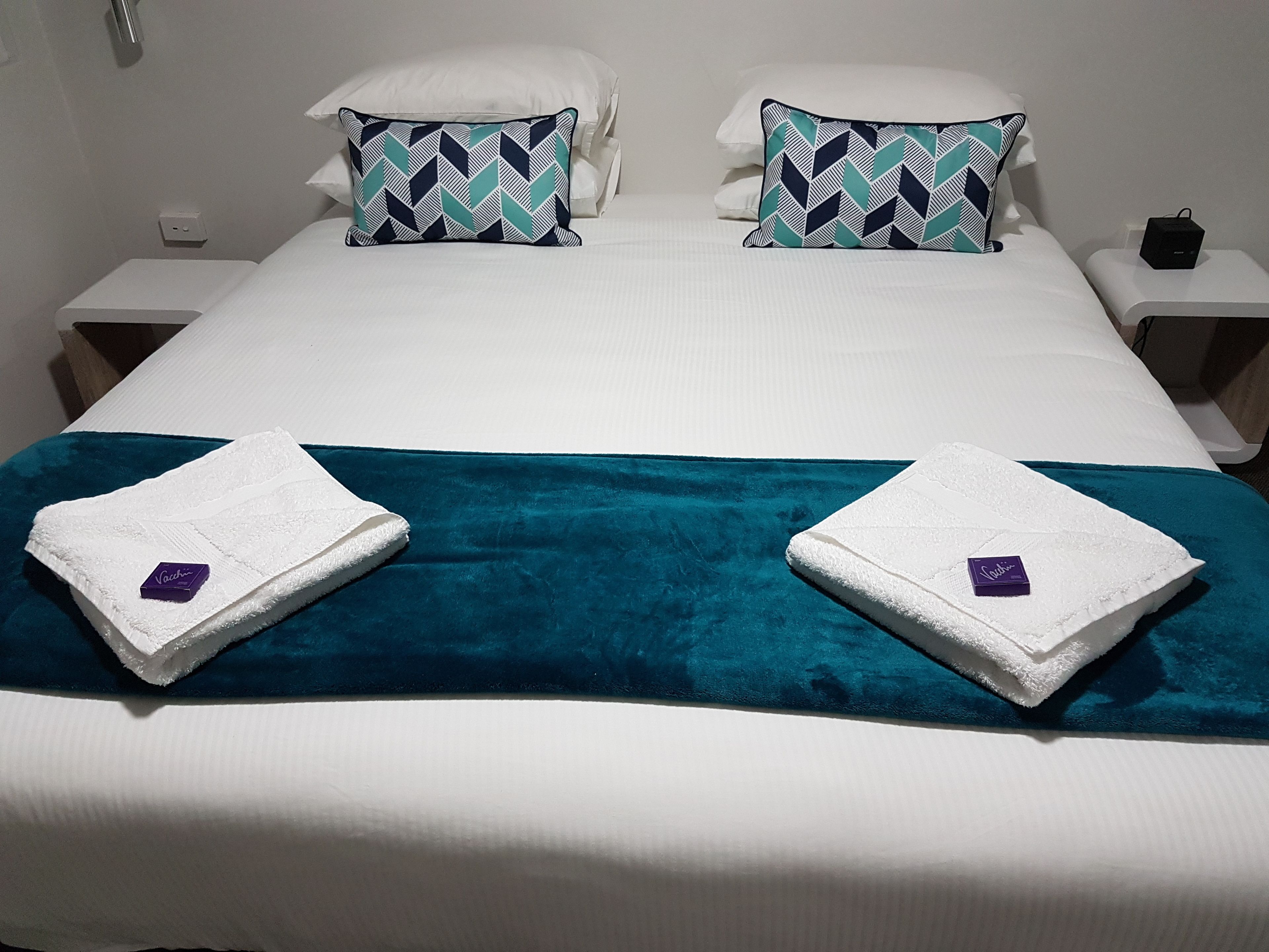 Una cama bien tendida con un plaid verde azulado y dos toallas con un logotipo púrpura.