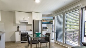 A modern kitchen with white cabinets, a stainless steel refrigerator, and a glass dining table with two chairs. There is a large window with blinds and a painting of a coastal scene. - Hamilton on Beaumont (Islington)