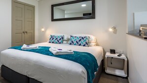 A neatly made bed with a teal blanket and white bedding, a bedside table with a lamp, and a door in the background. - Hamilton on Beaumont (Islington)
