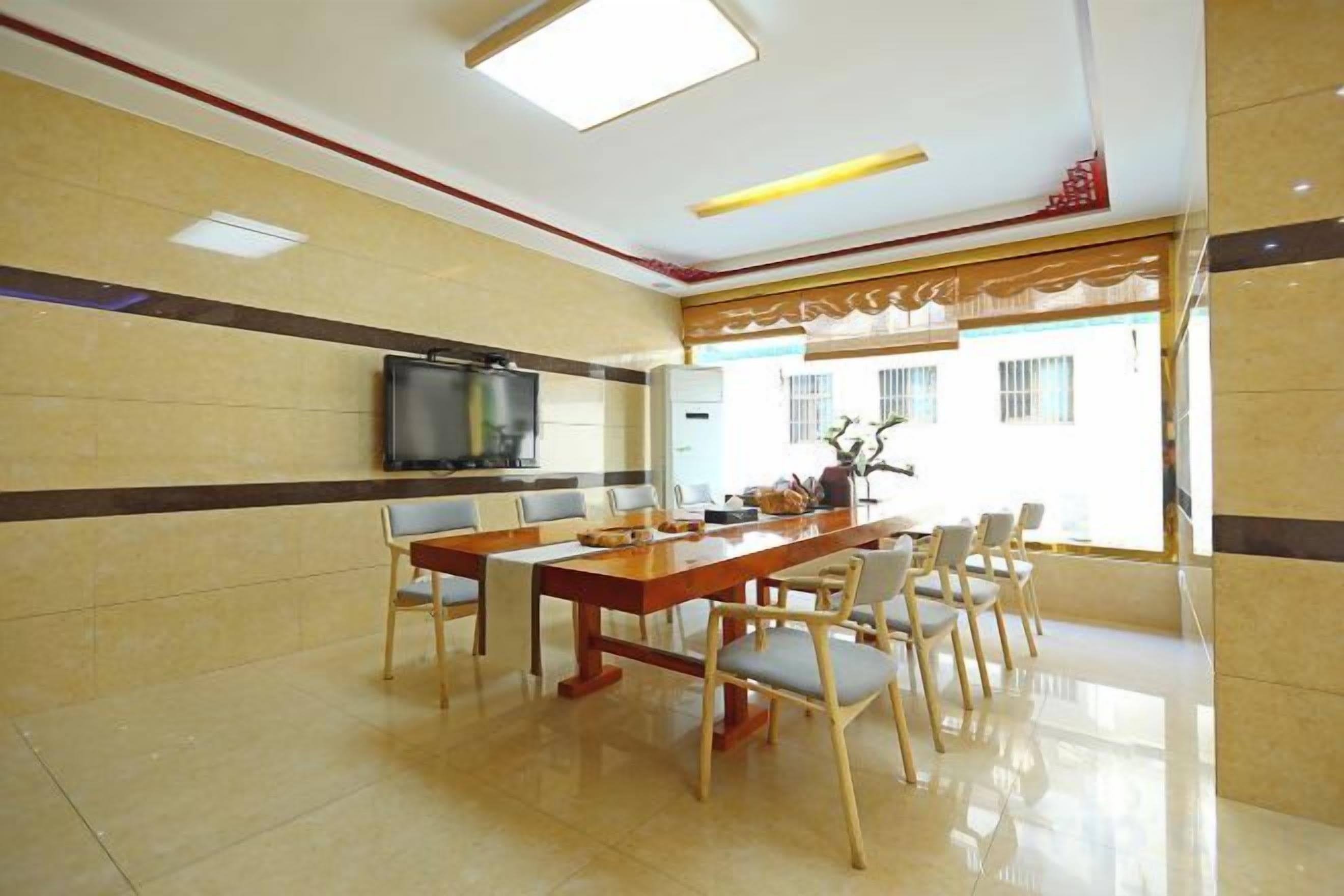 Comedor de hotel con una larga mesa de madera, varias sillas, un televisor de pantalla plana y vistas al exterior.