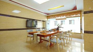 Comedor de hotel con una larga mesa de madera, varias sillas, un televisor de pantalla plana y vistas al exterior.