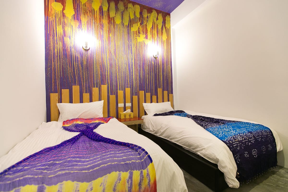 Quarto de hotel com duas camas, cada uma com edredom branco e manta colorida. Possui cabeceira de madeira com ripas verticais e luminária fixada na parede.